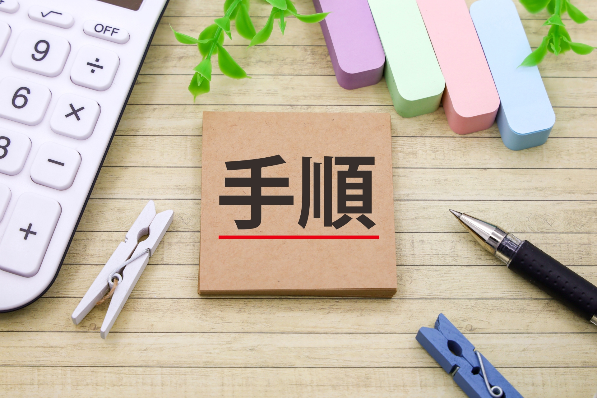 「手順」という文字が書かれたノートが、電卓や文房具と一緒に木製の机に置かれている。仕事や作業の順序やステップを計画する様子。