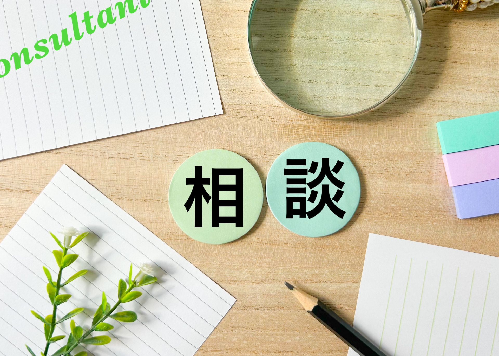 「相談」と書かれた2つの丸い紙が、ノートや文房具と共に置かれている。専門家への相談や、話し合いを通じて解決策を見つけることを表現している。